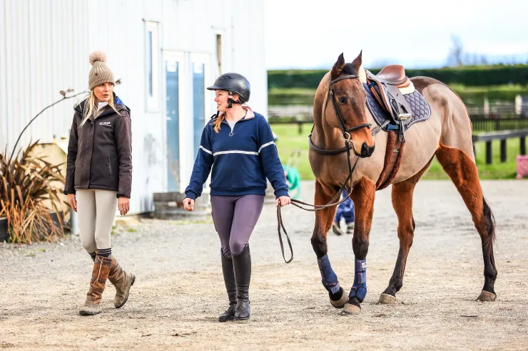 Woman leading a saddled horse beside another woman in a stable yard.