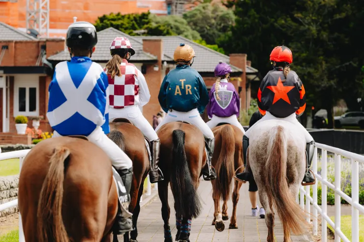 Jockeys in colourful silks riding horses along a track beside railings.